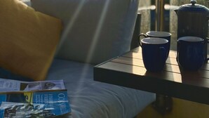 A balcony with a sofa, a table with two blue cups and a coffee pot, and a magazine.