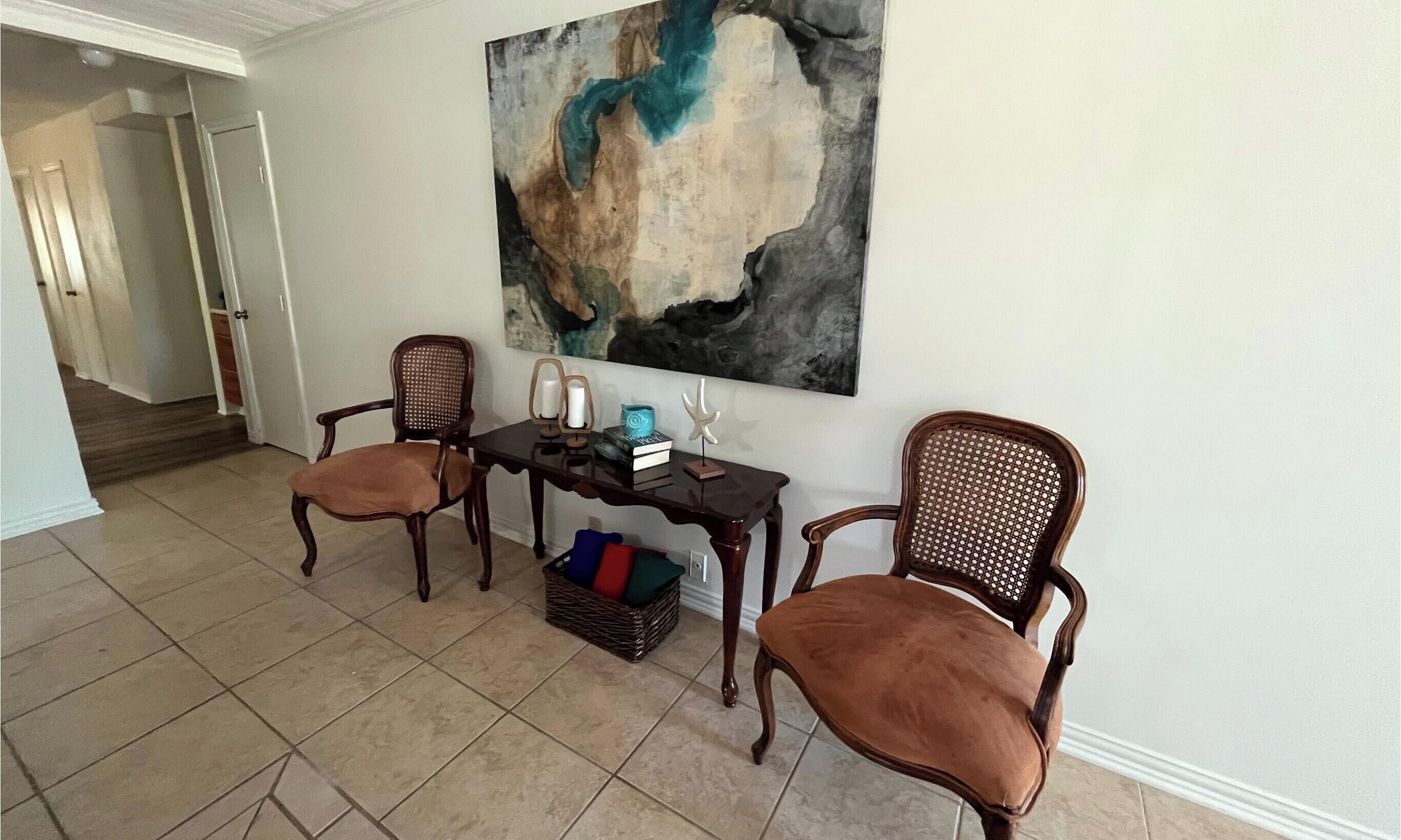 Ein Zimmer mit zwei Holzsesseln, einem kleinen Tisch mit Kerze, einem Buch und einem dekorativen Gegenstand. An der Wand hängt ein großes abstraktes Gemälde.