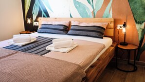 Un lit bien fait, avec une literie à rayures et deux serviettes, une tête de lit en bois, une table de chevet avec une lampe, et un mur orné d’une grande fresque représentant des plantes.