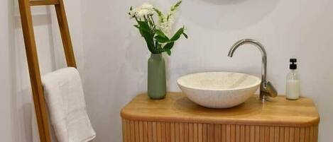 A bathroom with a wooden vanity, a round mirror, a white sink, a green vase with flowers, and a towel on a wooden ladder.