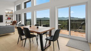 A spacious living area with a wooden dining table and chairs, a grey sofa, and a bookshelf. Large windows offer a view of a mountainous landscape.