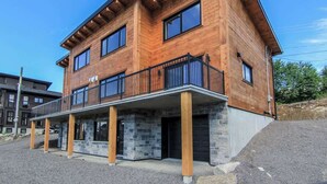 A two-story wooden house with a balcony, stone foundation, and a clear blue sky. - Olympe-215 | Tavata Chalets | Valinouët (Saint-David-de-Falardeau)