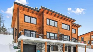 A modern multi-story wooden house with a stone foundation, large windows, and a covered front porch. - Olympe-215 | Tavata Chalets | Valinouët (Saint-David-de-Falardeau)