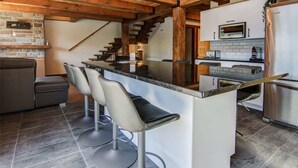 A kitchen with a central island, bar stools, a microwave, a refrigerator, and a staircase leading to another level. - Olympe-215 | Tavata Chalets | Valinouët (Saint-David-de-Falardeau)