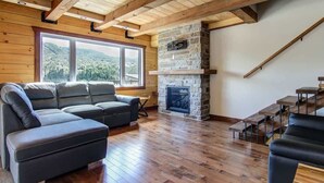 A cozy living room with a stone fireplace, wooden beams, and a large window overlooking a scenic landscape. - Olympe-215 | Tavata Chalets | Valinouët (Saint-David-de-Falardeau)