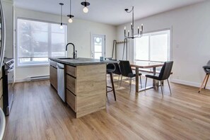 A modern kitchen with a central island, wooden cabinets, and a dining area with a table and chairs. - Remontée | Tavata Chalets | Valinouët (Saint-David-de-Falardeau)