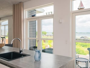 Eine Küche mit Spüle, einer blauen Karaffe und großen Fenstern mit Blick auf Grünflächen und ein Gewässer.