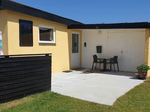 Il s’agit d’un bâtiment d’un seul étage, à la façade jaune et à la porte de garage blanche. Un banc noir se trouve devant le bâtiment, et une petite table avec deux chaises est placée près de la porte de garage.