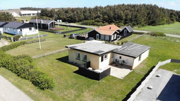 A residential area with two houses, one with a sloped roof and another with a flat roof, surrounded by a grassy field and a forest in the background. - 5 Star Holiday Home in Lokken (Lokken)