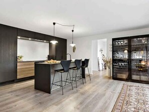 A modern kitchen with a central island, bar stools, pendant lighting, and a wine display cabinet. - 5 Star Holiday Home in Skagen (Skagen)