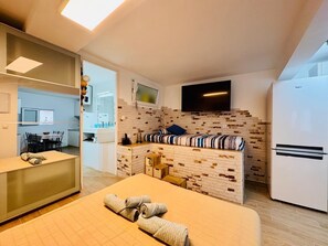 A compact hotel room with a kitchenette, a bed with rolled towels, a TV, and a refrigerator.