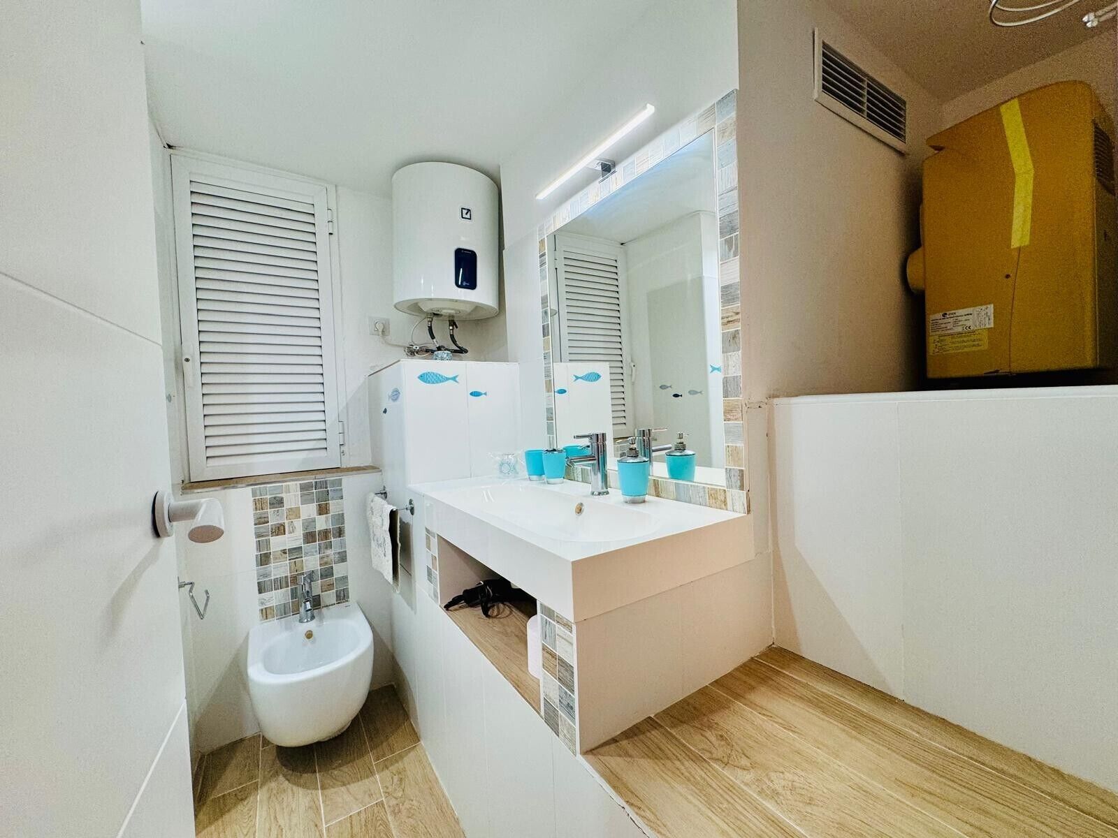Un bany amb un lavabo, mirall, vàter i una finestra amb persianes.