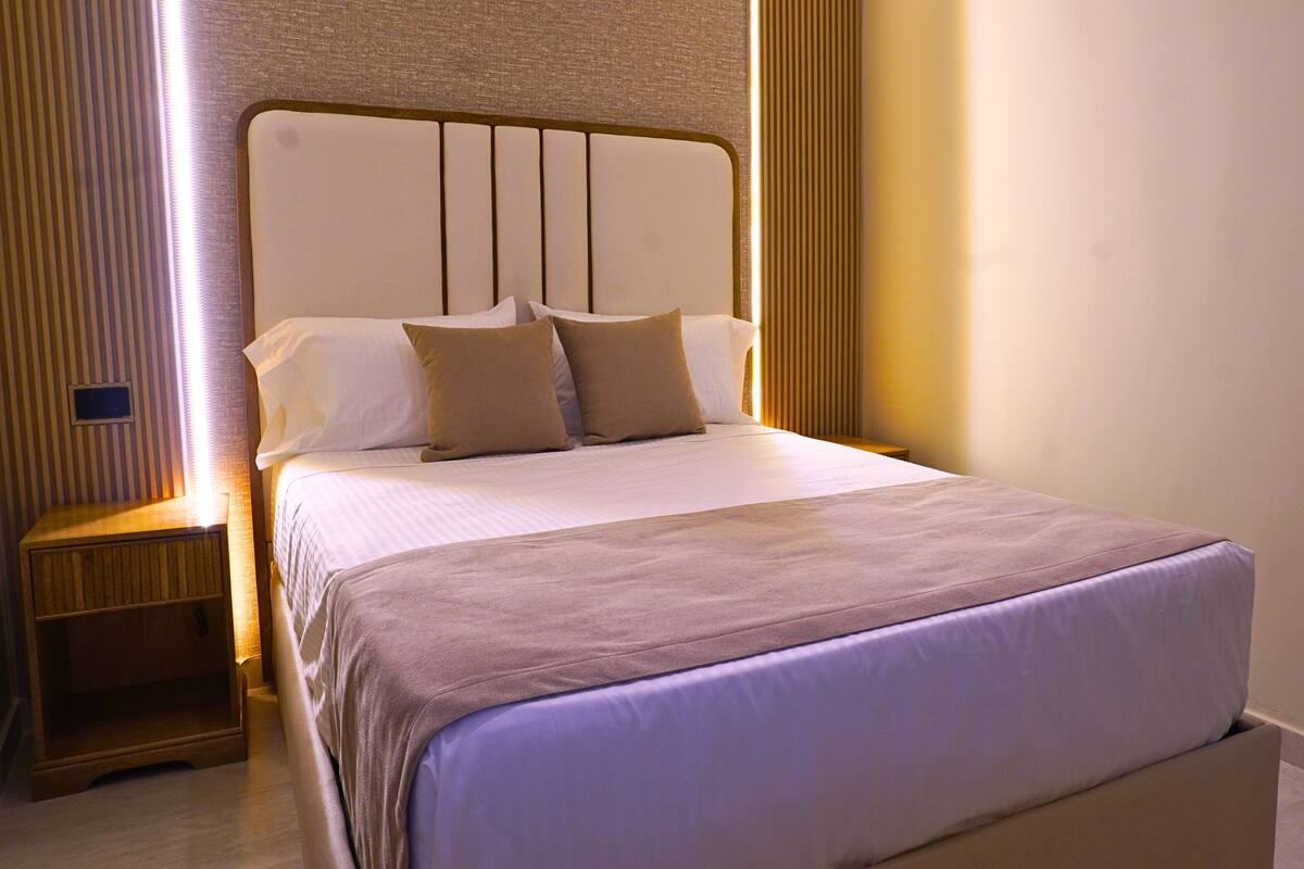 Un lit bien fait, avec une tête de lit, une table de chevet et un luminaire fixé au mur.