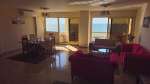 Un vaste salon comprenant un coin repas, un canapé rouge et une vue sur la mer grâce à de grandes fenêtres.