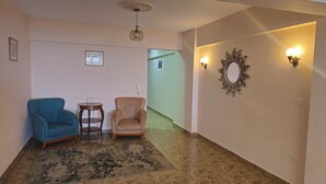 Un couloir avec un tapis à motifs, un fauteuil bleu, un fauteuil beige, une petite table avec un téléphone et un miroir décoratif.