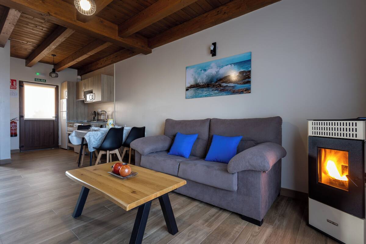 A living room with a sofa, a table, a fireplace, and a kitchen in the background.