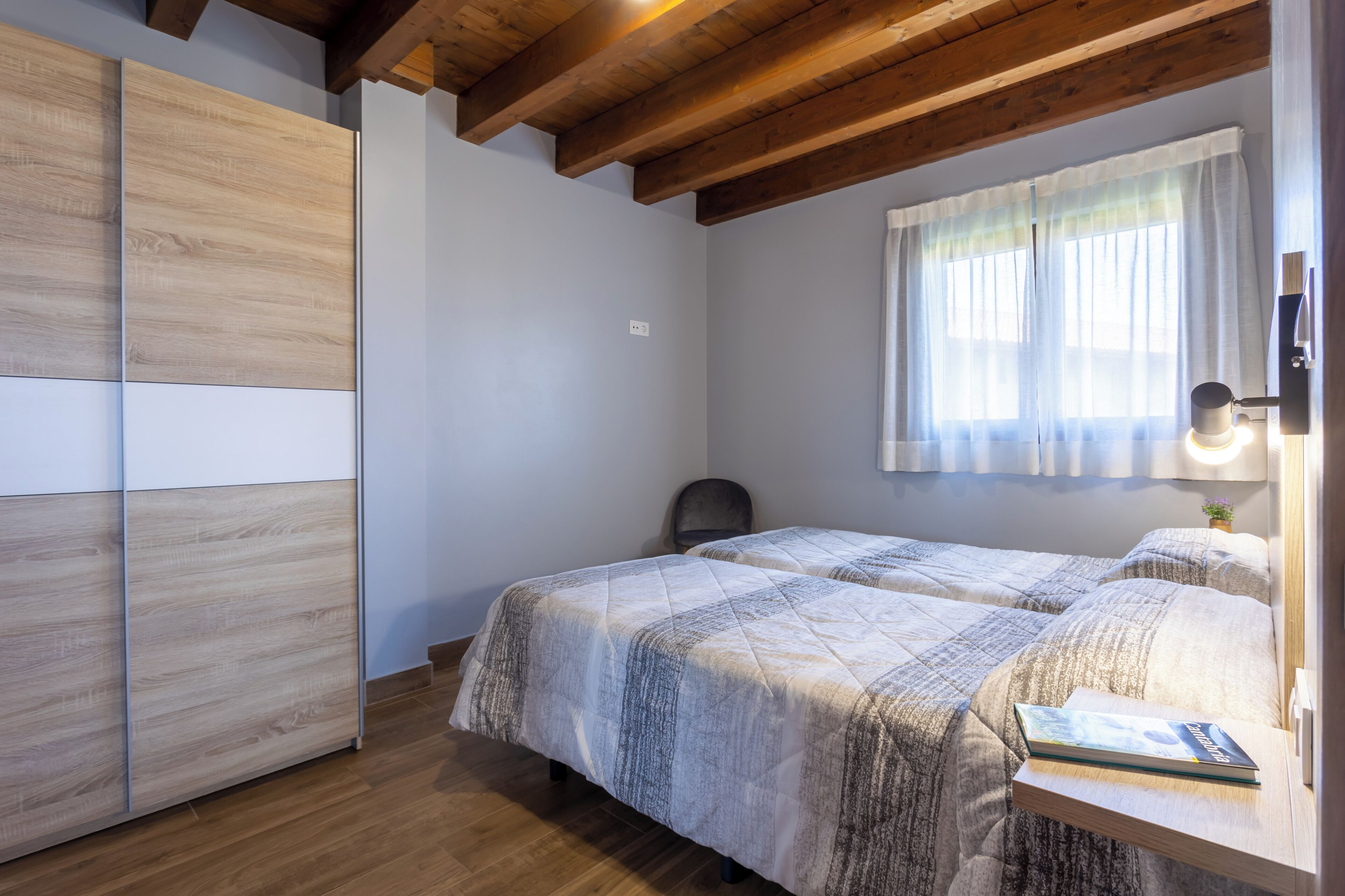 A bedroom with two beds, wooden flooring, a window with curtains, and a bedside lamp.
