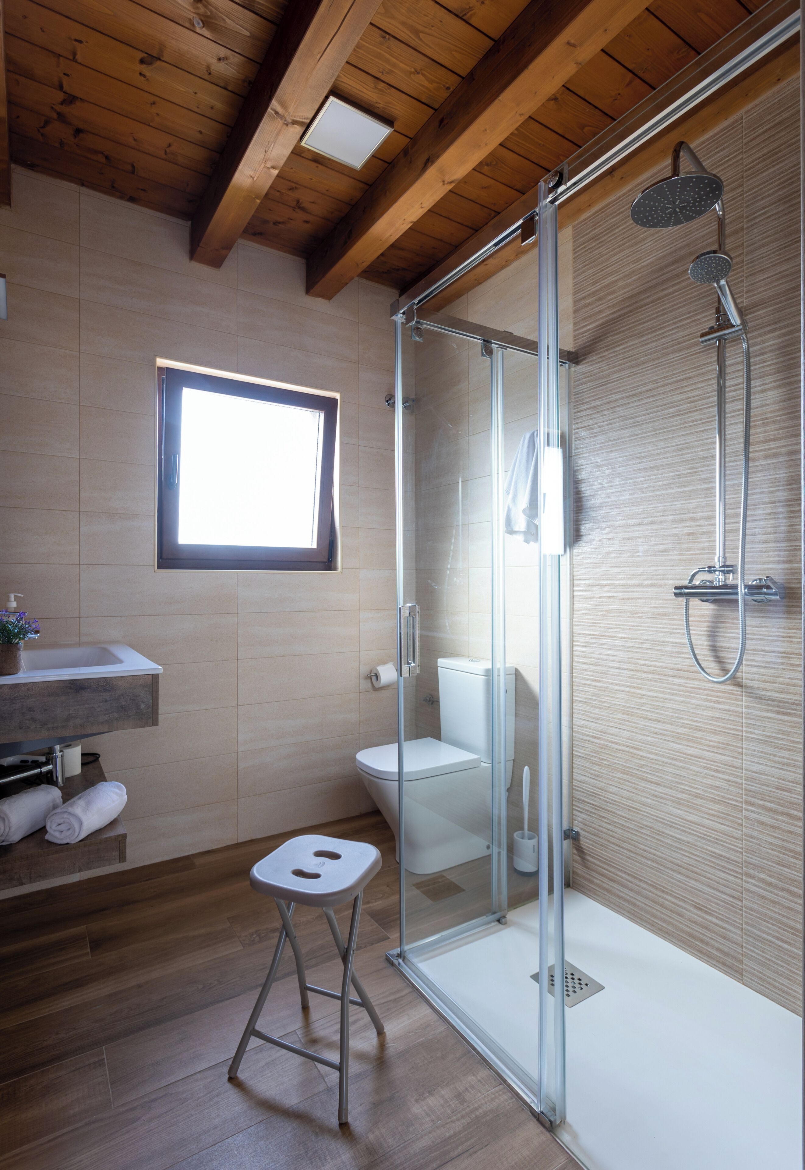 Un baño moderno con ducha envuelta en vidrio, techo de madera, ventana y un taburete pequeño.