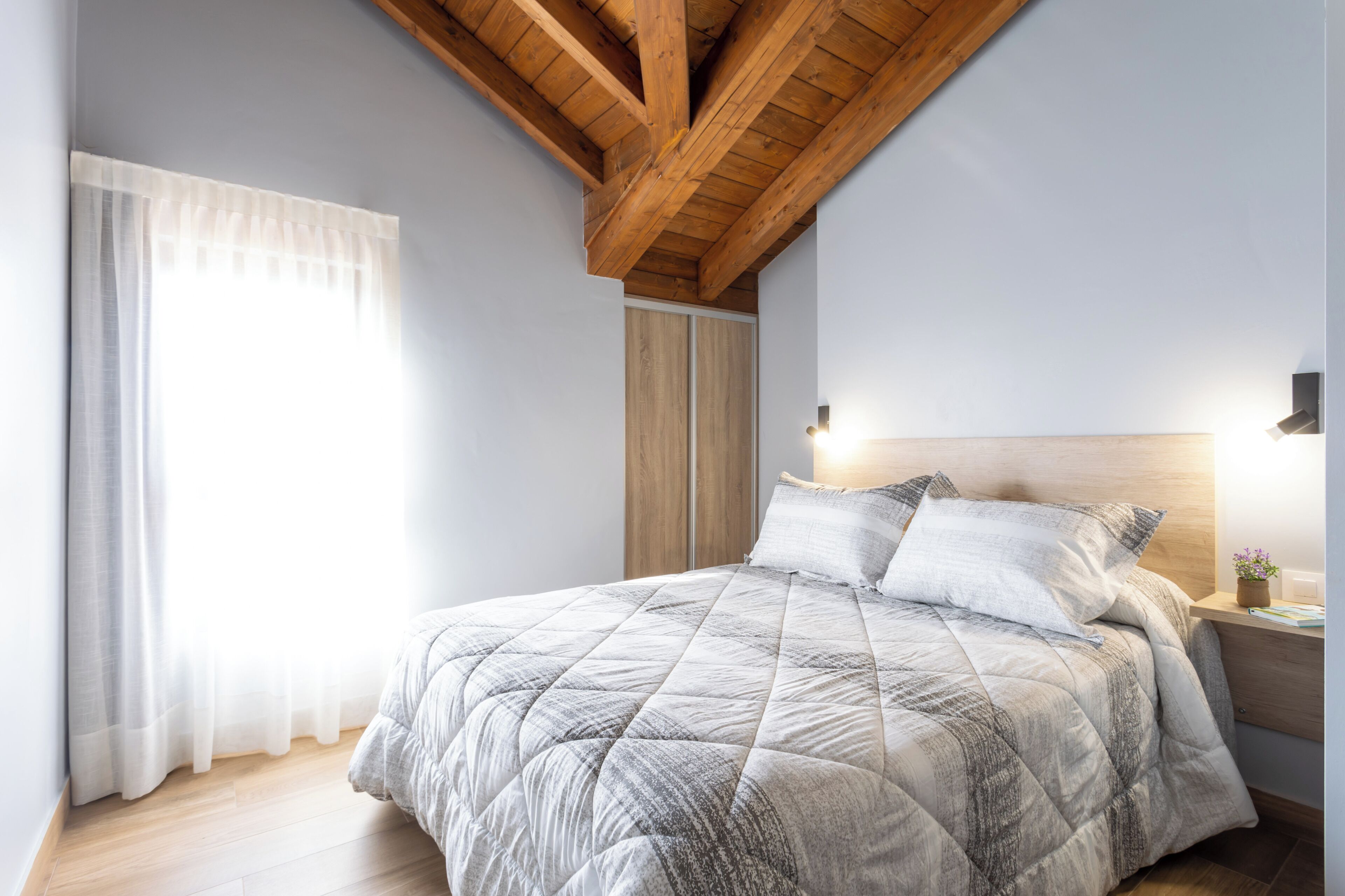 A bedroom with a wooden ceiling, a bed with a quilted bedspread, a nightstand with a lamp, and a window with sheer curtains.