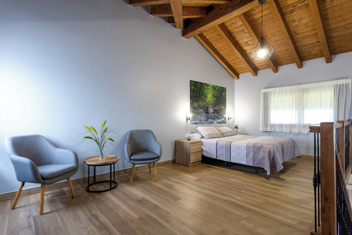 A bedroom with a bed, two chairs, a small table with a plant, a nightstand, and a large window.