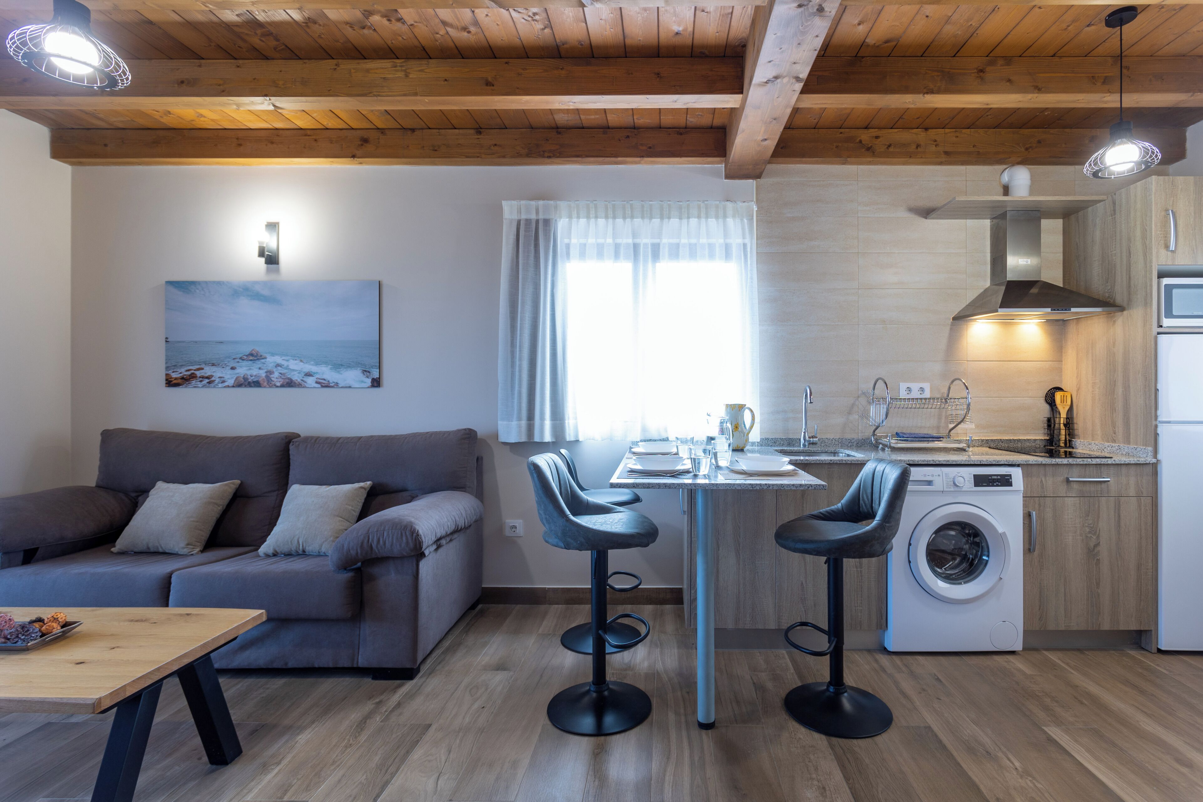 A compact living space with a sofa, two bar stools, a washing machine, a kitchen area, and a dining table.