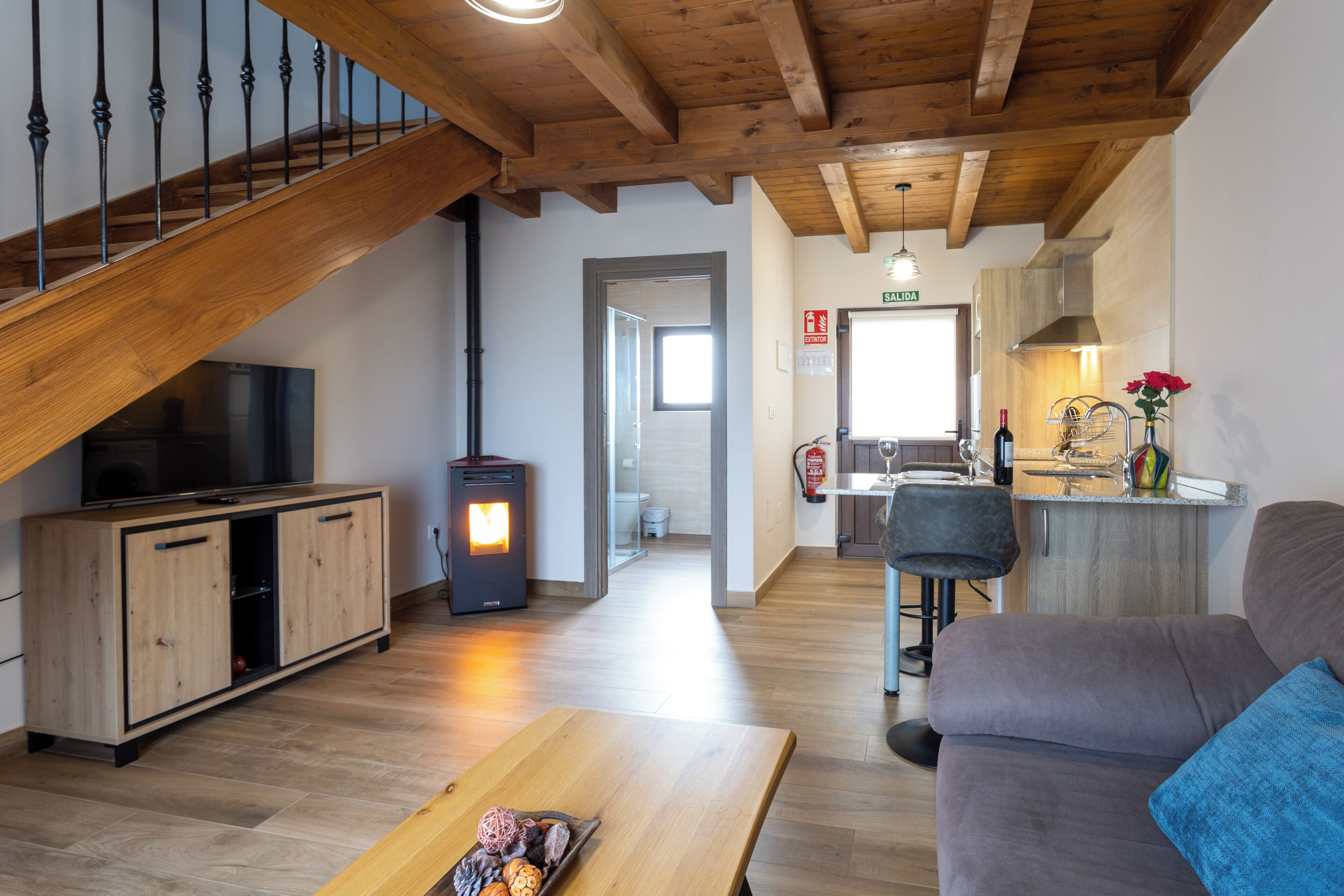 A modern living space with a wooden staircase, a television on a wooden cabinet, a fireplace, a sofa, and a kitchen area.