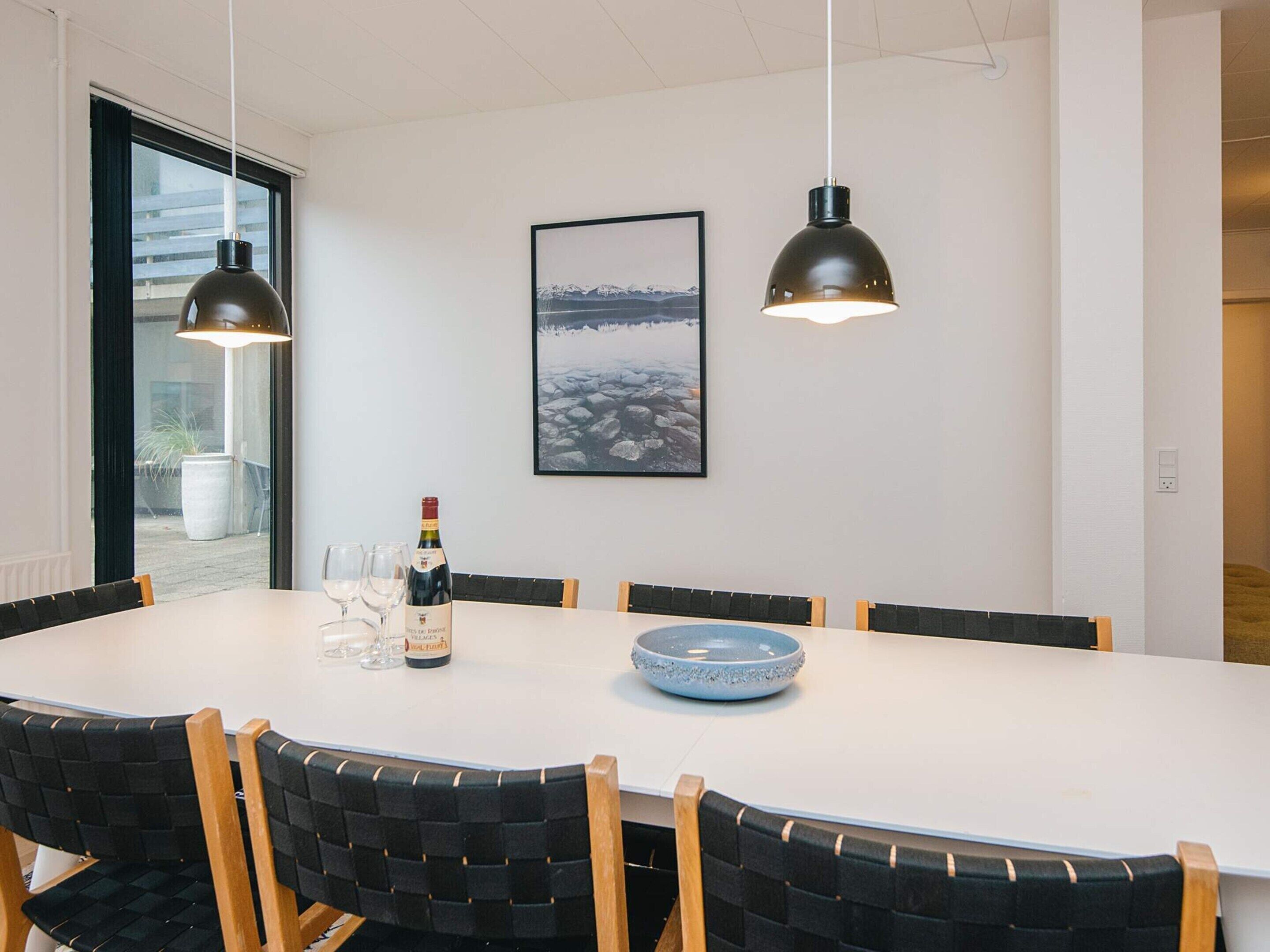 A dining area with a white table, black upholstered chairs, two pendant lights, a bottle of wine, and a framed picture on the wall.