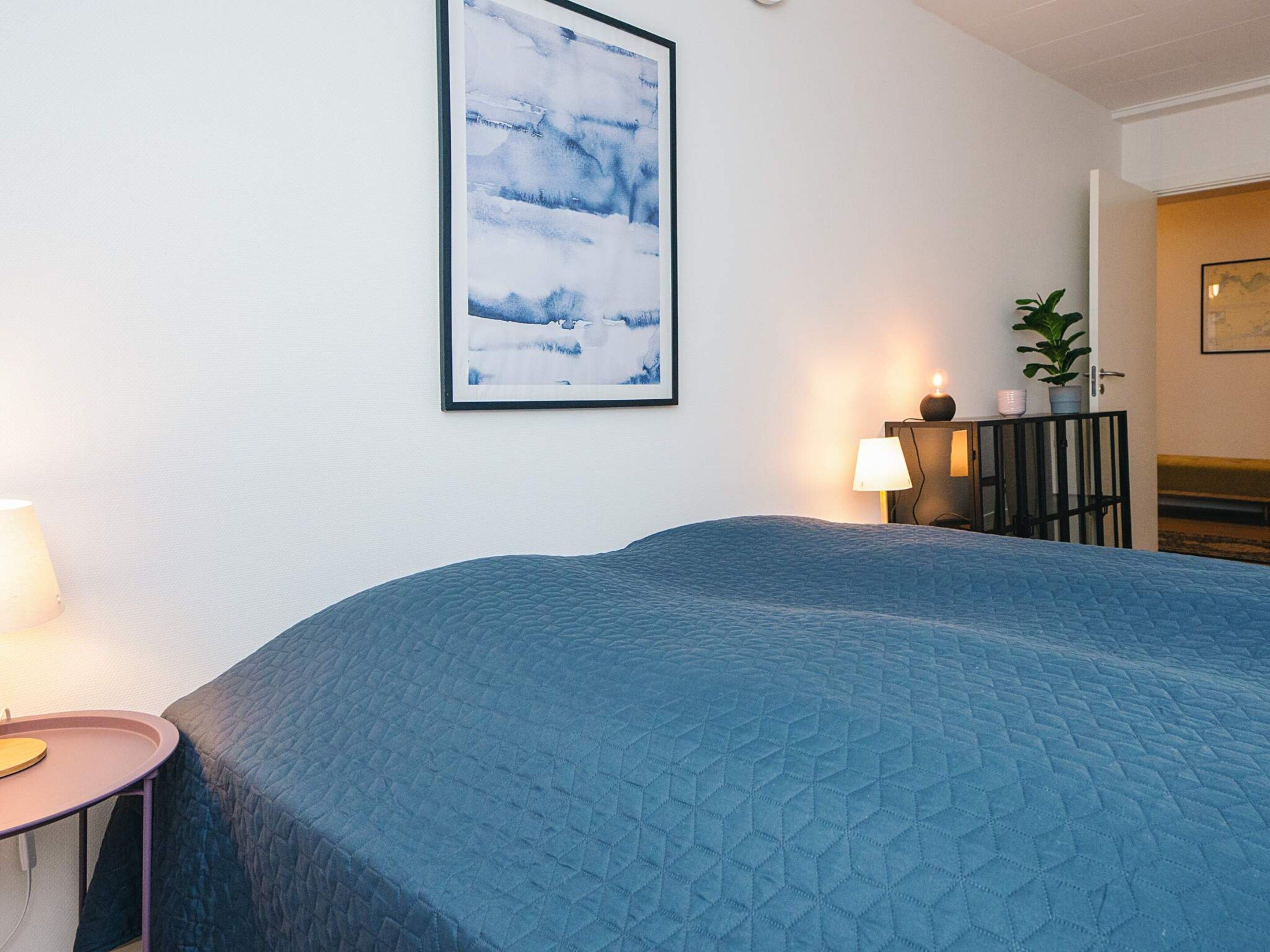 A bedroom with a large bed, a bedside table with a lamp, a framed picture on the wall, and a potted plant on a cabinet.