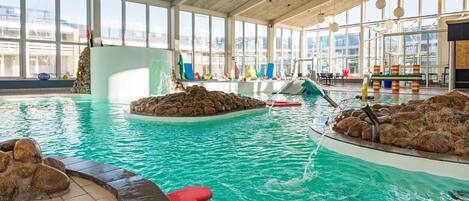 Piscina interna com água cristalina, formações rochosas e vista da cidade através de amplas janelas.