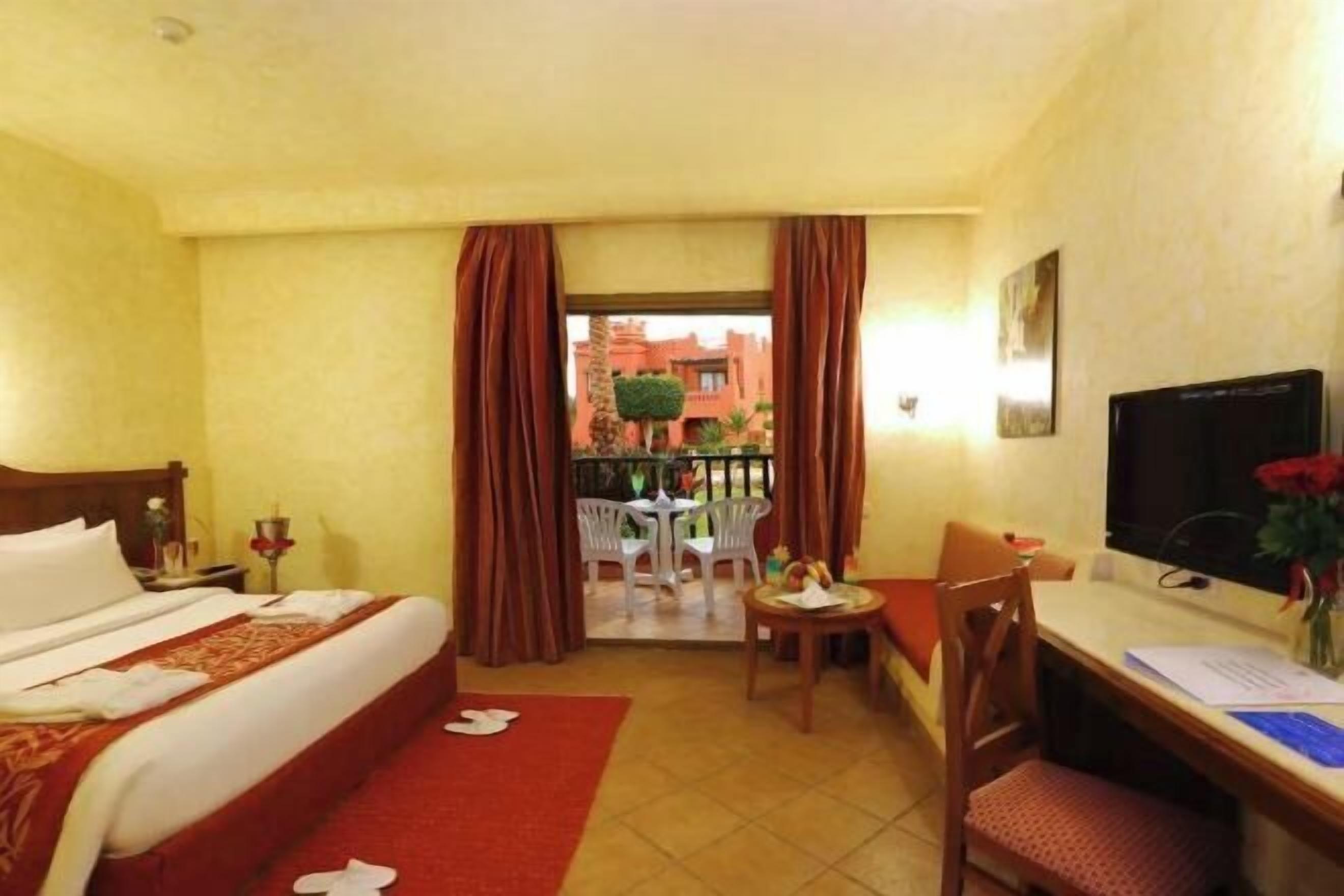 Habitación de hotel con cama, escritorio, silla, televisión y balcón con vista a edificios.