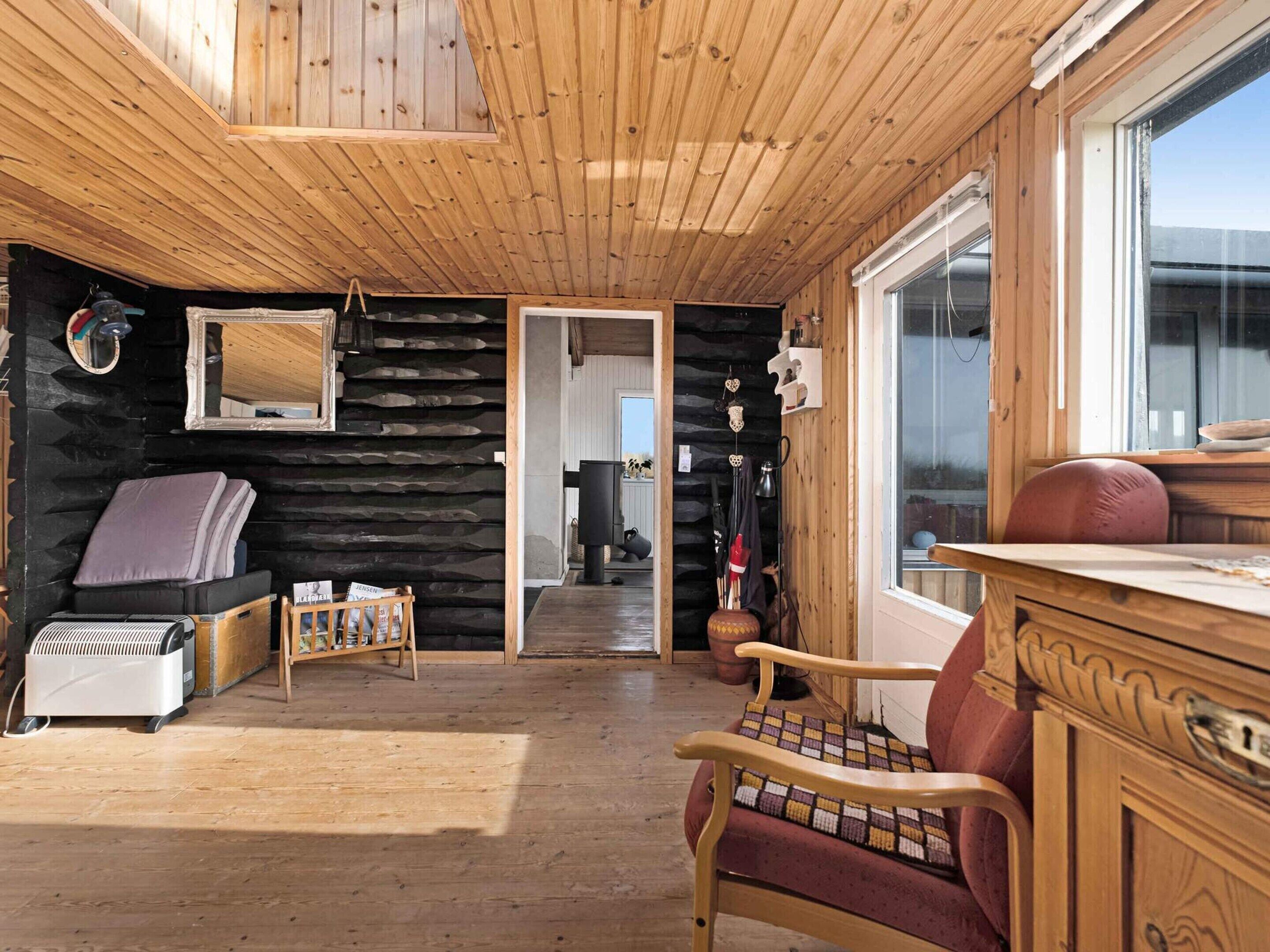 A cozy cabin interior with a wooden ceiling, a stone wall, a bed, a chair, and a window.