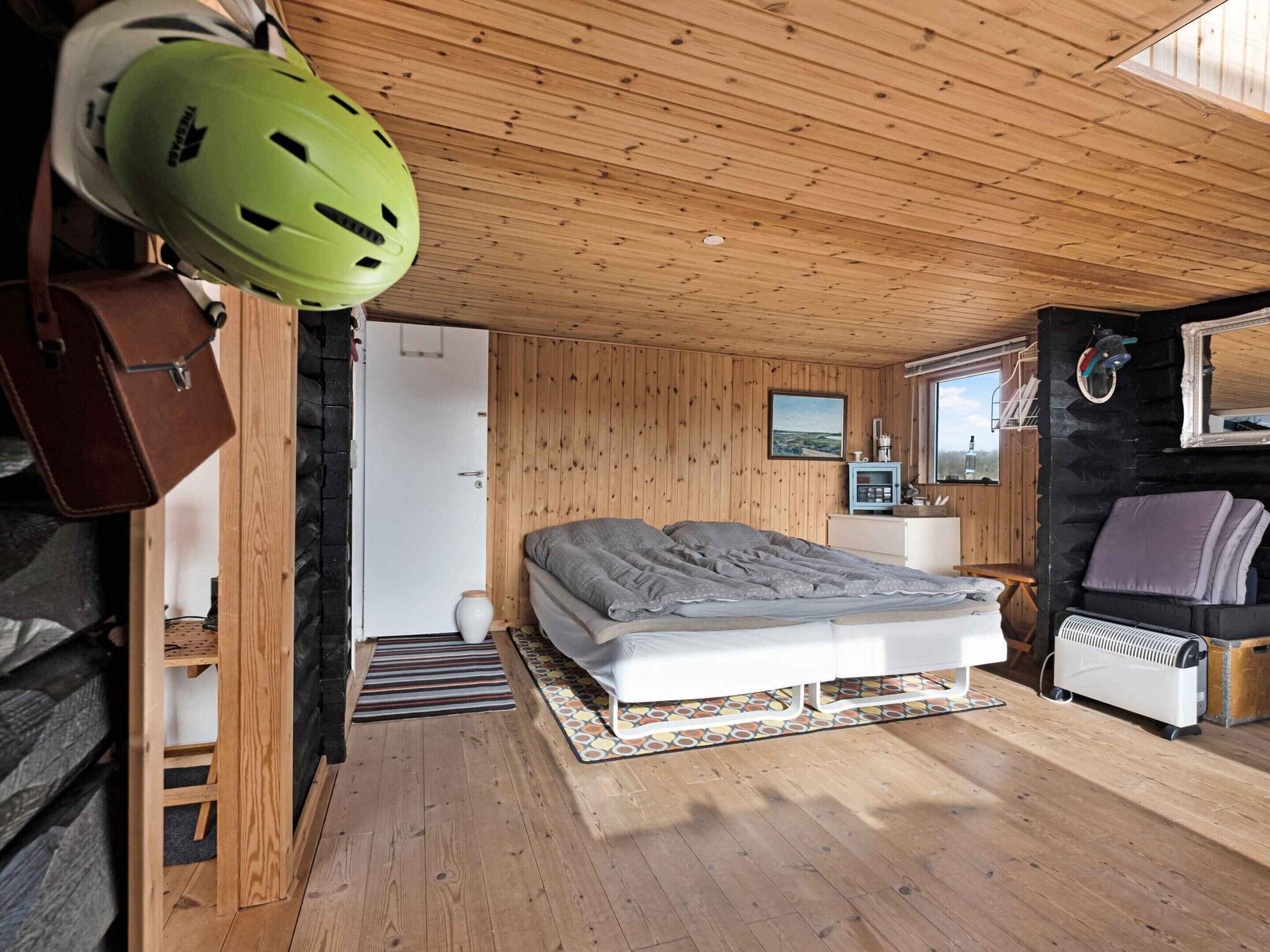 A cozy wooden cabin bedroom with a bed, a small bedside table, a heater, and a helmet hanging on the wall.
