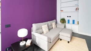 Ein modernes Wohnzimmer mit einer violetten Wand, einem grauen Ecksofa, einem kleinen Beistelltisch mit Lampe und einer weißen Regaleinheit mit Büchern und Dekoration.