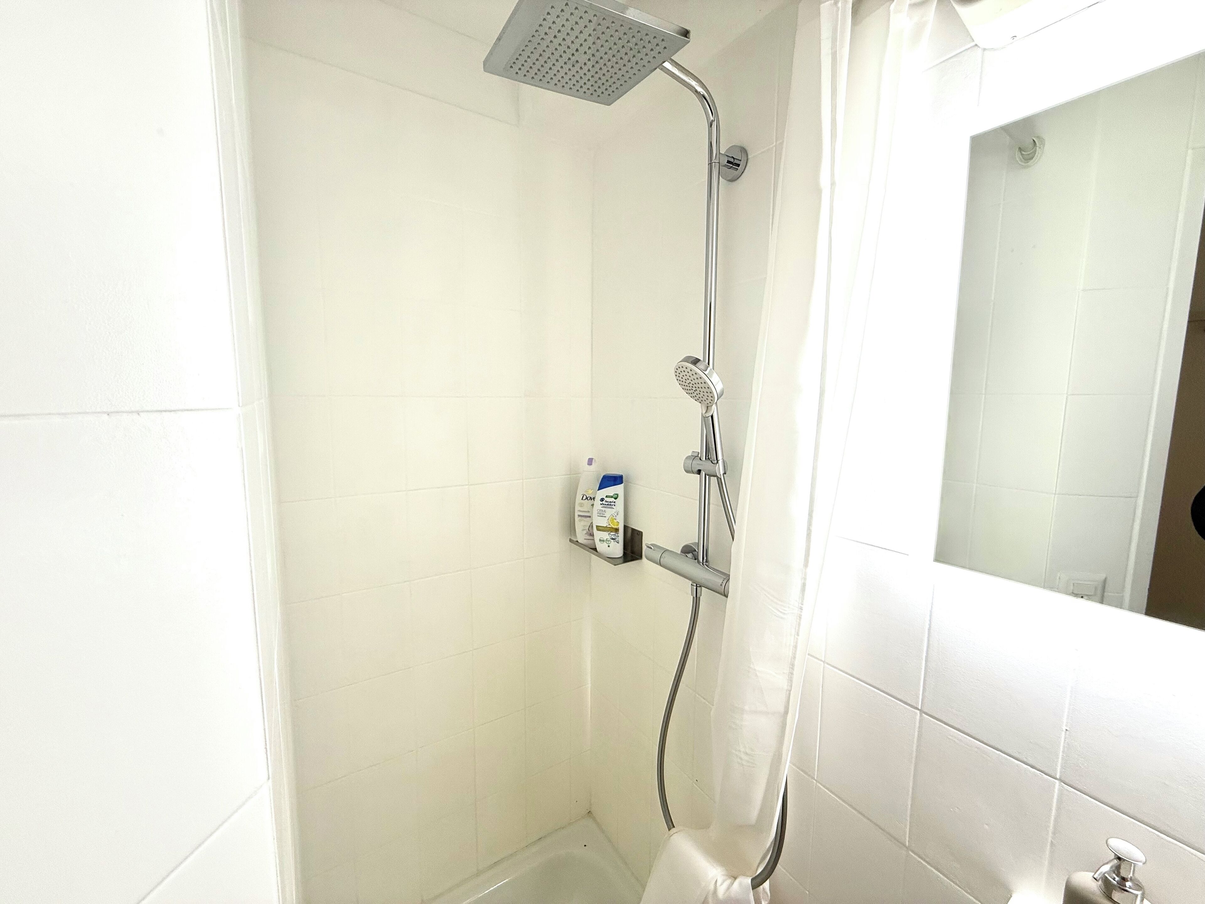 A small, white-tiled bathroom with a shower, a handheld showerhead, a wall-mounted soap dispenser, and a mirror.