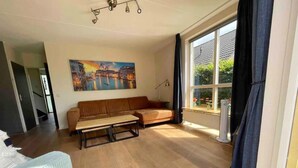 A living room with a sofa, a coffee table, a painting, and a window with curtains. - Family Bungalow With Private Garden (Ewijk)