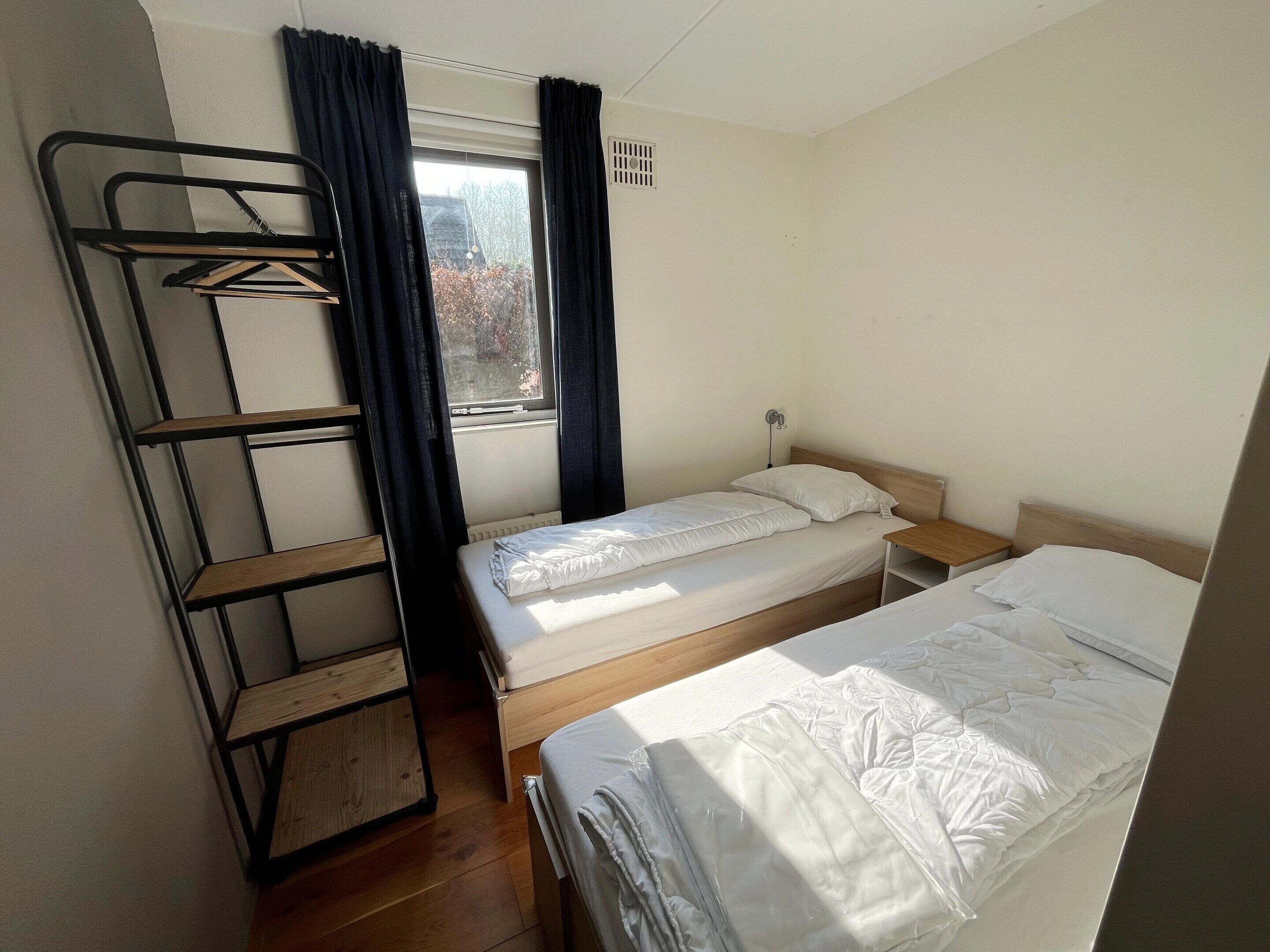 A dormitory room with bunk beds, a window with curtains, a radiator, and a wooden shelf unit.