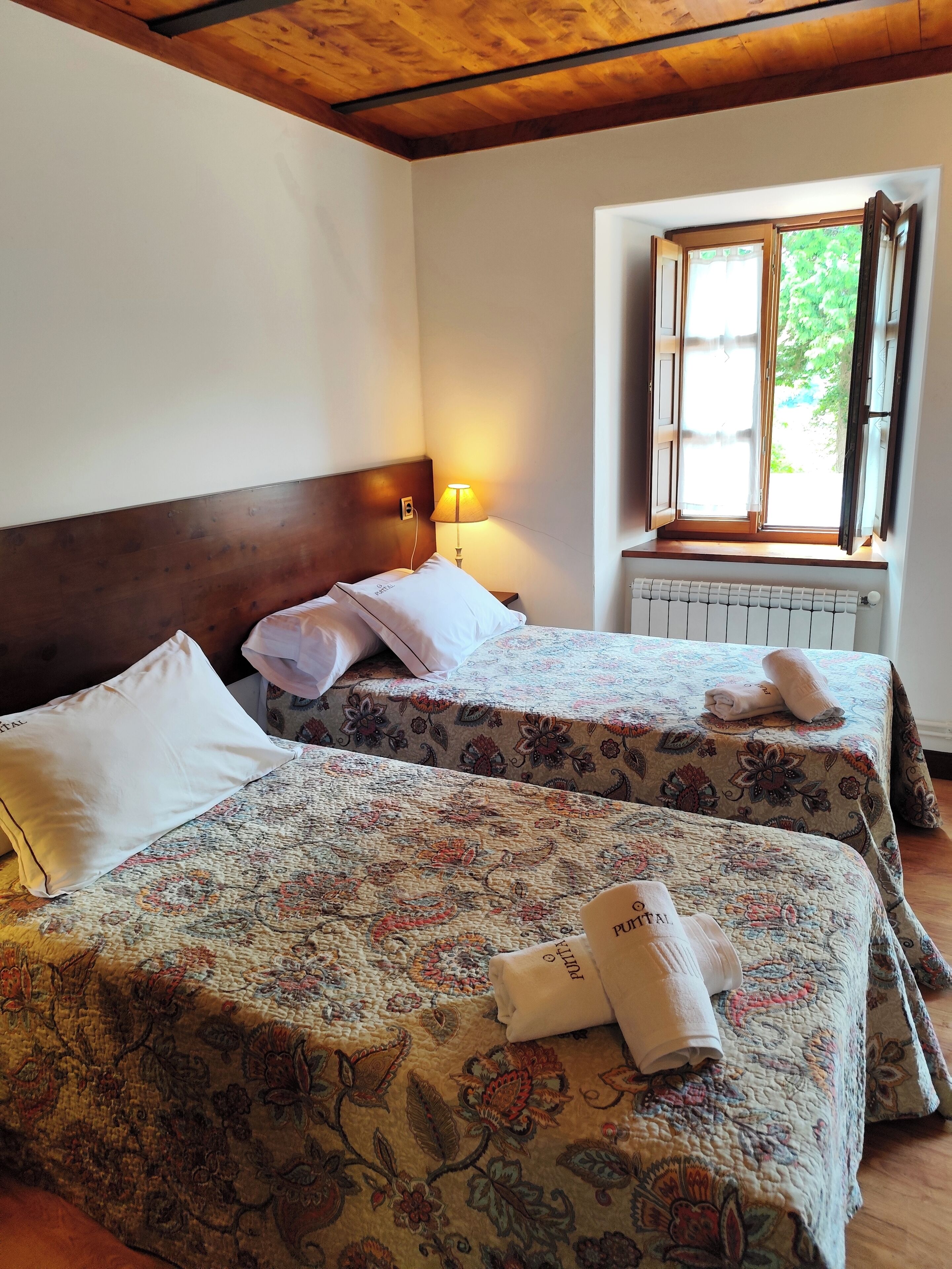 A hotel room with two beds, each with floral-patterned bedding and white pillows. There is a window with open shutters, a wall-mounted radiator, and a lamp on the nightstand.