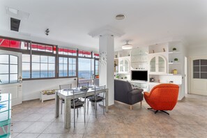 Un espacio habitable amplio con una mesa de comedor, un sofá, un televisor y vistas al mar a través de grandes ventanales.