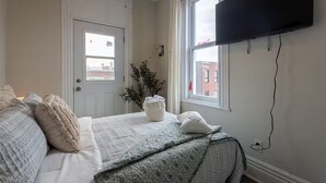 A bedroom with a bed, a television mounted on the wall, a door, and a window with a view of buildings outside. - Confortable Home on Notre Dame & near Downtown (Montreal)