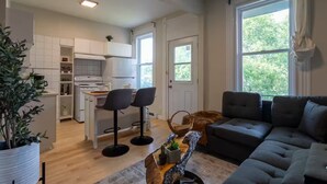 A modern kitchen with white cabinets, a dining area with two black bar stools, a grey sofa, and a large window offering a view of greenery. - Confortable Home on Notre Dame & near Downtown (Montreal)
