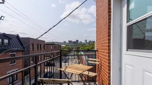 A balcony with a wooden table and chairs, overlooking a cityscape with buildings and a street. - Confortable Home on Notre Dame & near Downtown (Montreal)