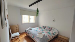 Un dormitorio con cama, escritorio, silla, ventana y una planta colgante.