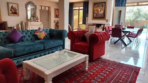 Een ruime woonkamer met een blauwe bank, een rode fauteuil en een glazen salontafel. Er is een eethoek met rode stoelen en een kamerbreed tapijt met een patroon.