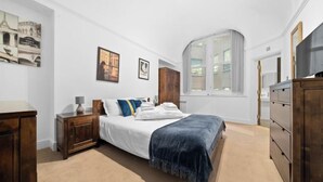 Une chambre à coucher comprenant un lit, une table de chevet, une commode, une fenêtre et des œuvres d’art murales.