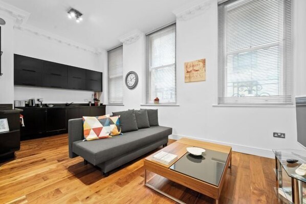 A modern living room with a sofa, coffee table, and kitchen area in the background. - Apartment 2, 48 Bishopsgate by City Living London (London)