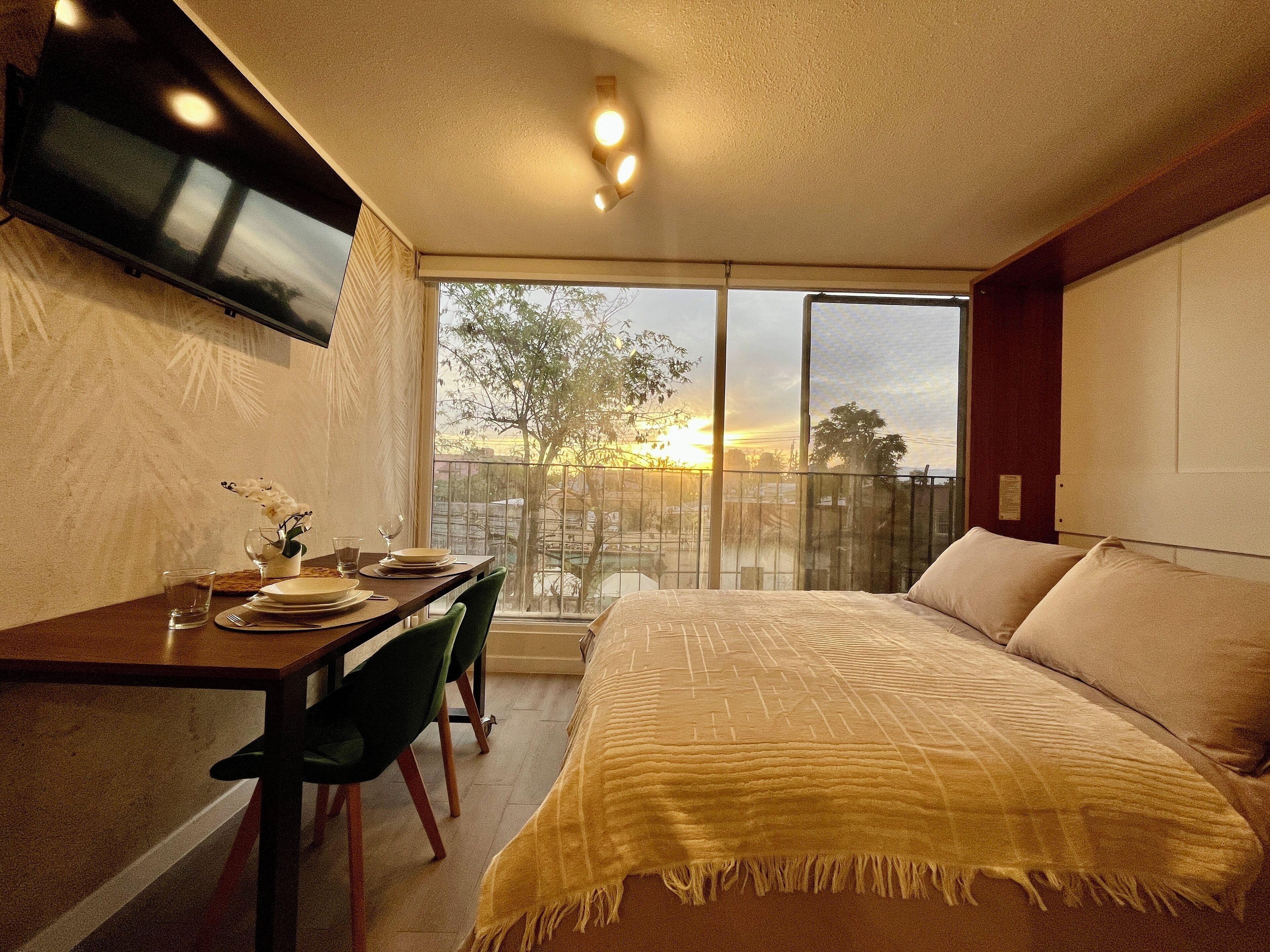 A bedroom with a bed, a desk with a chair, a TV mounted on the wall, and a large window with a view of the sunset.
