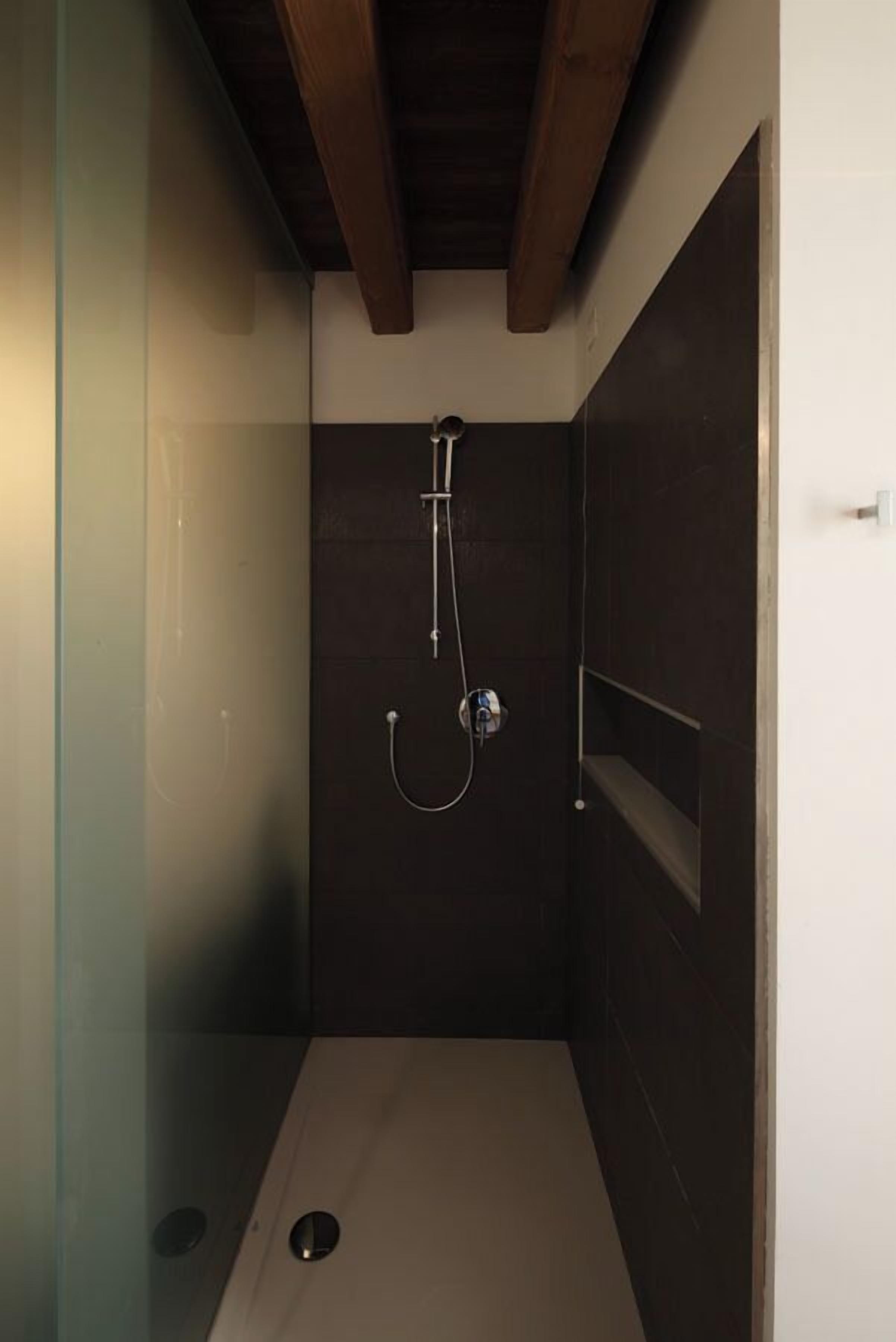 Een kleine, afgesloten douchecel met een donkere betegelde wand, een houten balk in het plafond en een glazen deur.