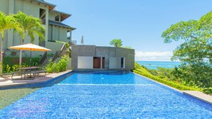 Une piscine avec vue sur l’océan, un patio couvert avec une table et des chaises, et un petit bâtiment doté d’un balcon.