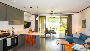 Une cuisine moderne dotée d’un îlot central, un coin repas avec une table et des chaises, et un salon avec un canapé et un ventilateur de plafond.