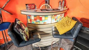 Un coin bar agrémenté d’un mur recouvert d’une mosaïque colorée, deux tabourets de bar noirs avec des coussins jaunes, une chaise et une petite table sur laquelle sont posées des bouteilles et un menu.